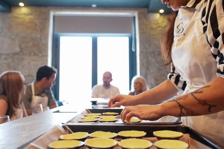 Woman preparing dough in tart molds with people seated in the background in a kitchen.