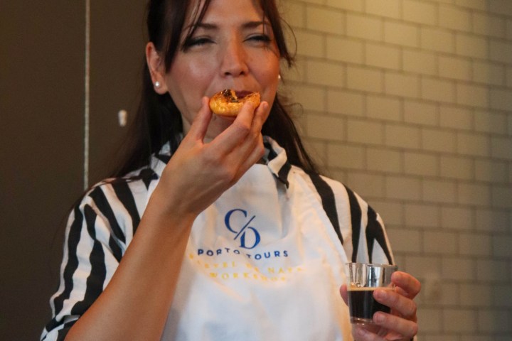 Woman enjoying pastry and holding a drink, wearing a striped shirt and apron in a tiled room.
