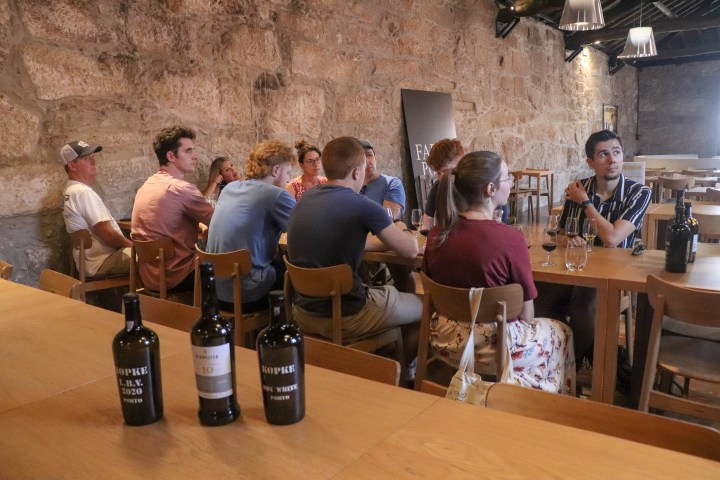 Group of people sit at a long table with wine bottles in a rustic, stone-walled room.