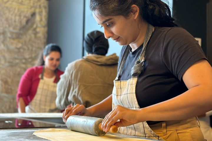 Woman in apron rolling dough with a pin in a kitchen with others in background.