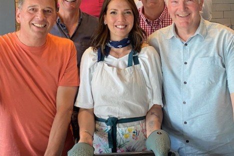 Seven people smiling with woman holding a tray of baked custards in a kitchen.