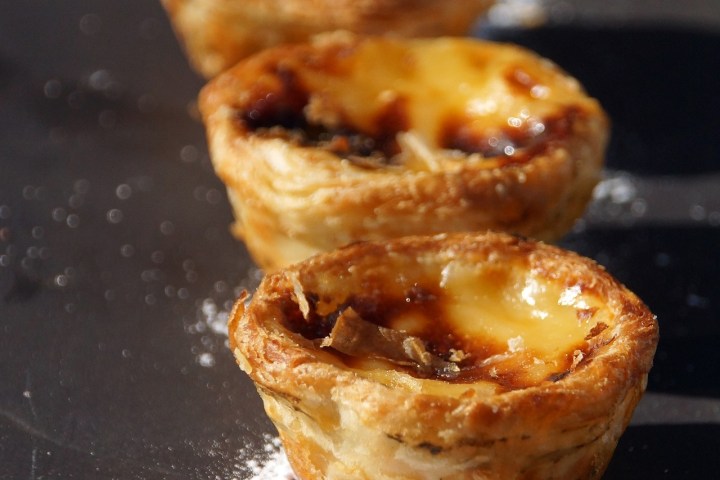 Four Portuguese egg tarts on a dark surface with powdered sugar.