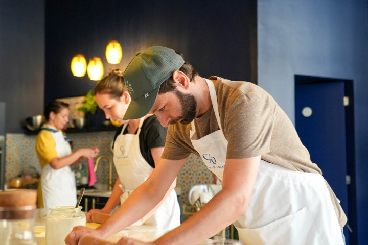 People wearing aprons, rolling dough in a kitchen with blue walls and warm lighting.
