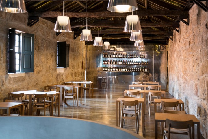 Rustic bar with wooden tables, chairs, pendant lights, and bottles on backlit shelves.