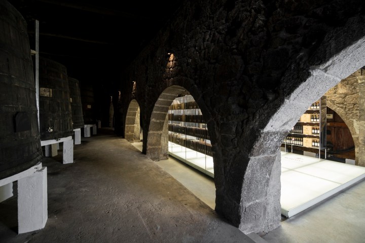 Stone arches in a dim wine cellar with large barrels and shelves of bottles.