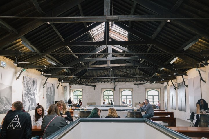 People sitting at tables in an industrial-style room with high ceiling and large windows.