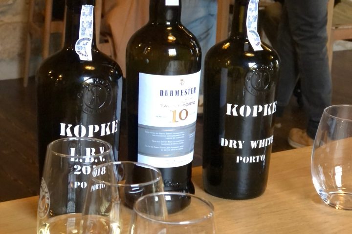 Three wine bottles with red pour spouts beside glasses of red and white wine on a wooden table.