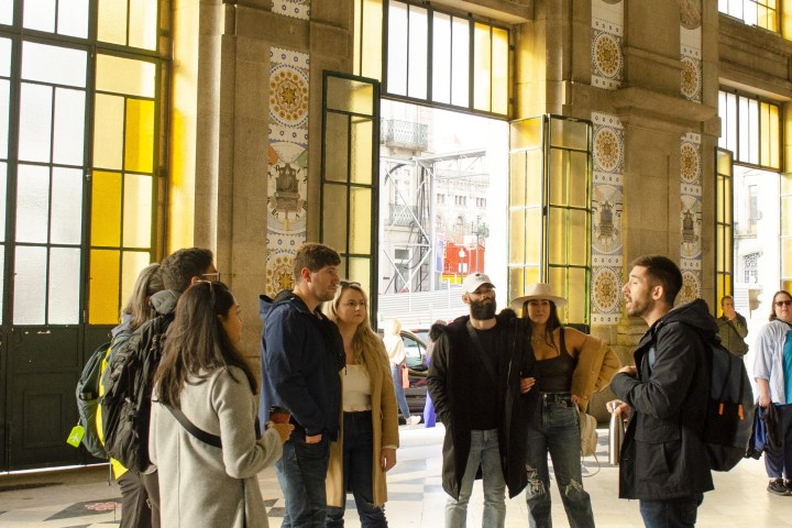 Group of people standing in a spacious, sunlit building with tall windows and decorative columns.