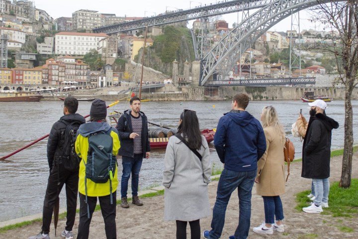 Group of people near a river, with a bridge and city in the background.