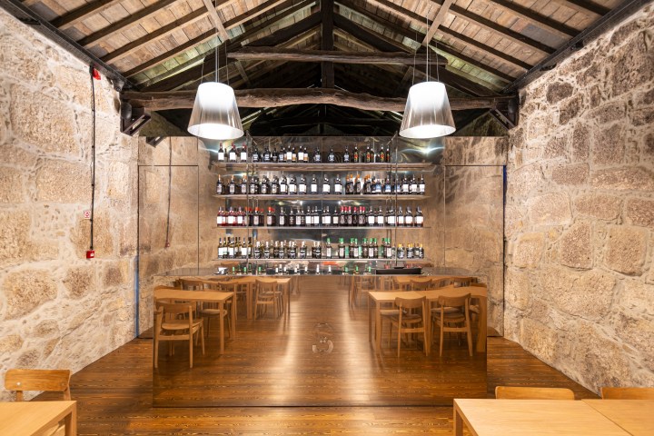 Rustic stone-walled room with wooden tables, chairs, and shelves of bottles, under dim pendant lights.