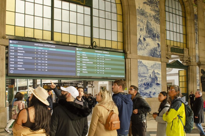 People in a train station looking at a departures board under a large clock and arched windows.